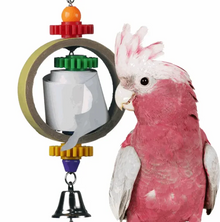A pink and white cockatoo standing beside a colorful bird toy with gears, a bell, and a hanging ring on a plain white background.